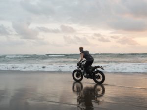 Side view of faceless male rider leaned forward while riding bike on wet sand near ocean with foamy waves under cloudy sky at sundown